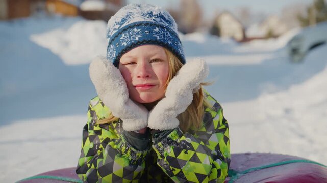 Young child smiles warmly in snowy winter scene. Wears blue hat and bright green jacket. Hands covered in white gloves, cheeks flushed from cold. Looks joyful, excited for snow fun