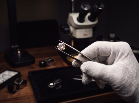 A hand in a white glove holds a brilliant diamond with tweezers, examining it under a microscope in a dark, sophisticated setting