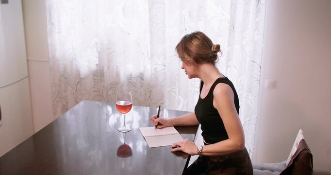 Home inspiration. Creative writing. Thoughtful woman working in notebook at table with orange drink in glass enjoying quiet routine.