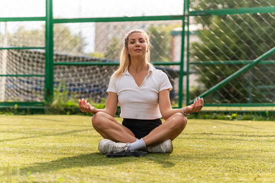 Calm woman meditating in a lotus pose on a green sports field with eyes closed. Yoga practice, mindfulness, and morning relaxation.