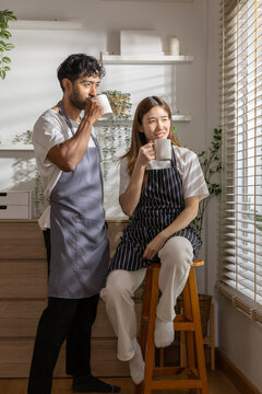 Happy young asian couple wearing apron enjoying morning coffee from white mug in bright kitchen near window, showing cheerful smile during peaceful weekend together. Vertical