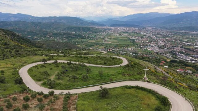 Drone flies over a winding road near Elbasan with a cross and city in the valley.