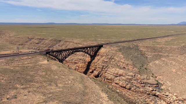 4K UHD drone flight capturing an industrial railroad bridge cutting through the remote Southwest plains. The heavy steel structure spans a sheer, rocky chasm under a bright, expansive sky.