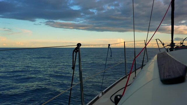 serene sailboat stationary on open sea at dusk from the deck of a sailboat looking out to sea. travel and adventure, nature and landscapes. blue ocean water, cloudy sky, wooden sailboat.