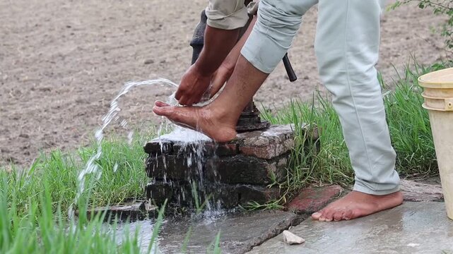 Close-up of an unrecognized rural Indian man washing his muddy bare feet with fresh flowing water from a traditional manual handpump outdoors.
