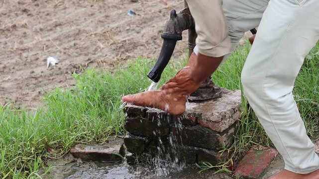 Close-up of an unrecognized rural Indian man washing his muddy bare feet with fresh flowing water from a traditional manual handpump outdoors.
