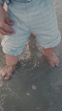 4K Vertical close up of a baby boy's feet standing in shallow sea water on a sandy beach.