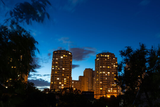 Urban district. High residential buildings in front of a dark blue evening summer sky with tree silhouettes. City at night