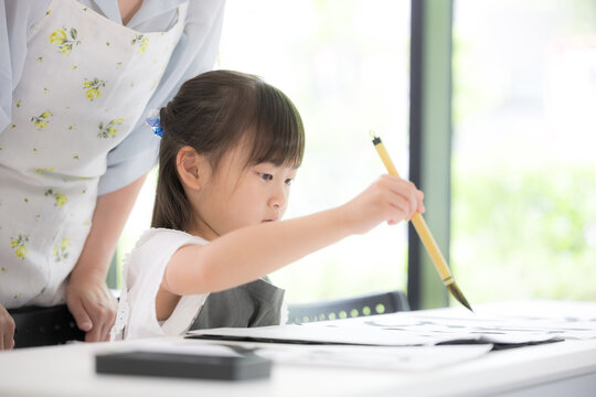 An elementary school girl concentrating on calligraphy as she writes characters with a brush; Japanese children learning calligraphy, taking instructions from their teacher with serious expressions