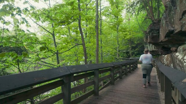 A woman walking on a boardwalk surrounded by rocks and trees while taking pictures on her phone.