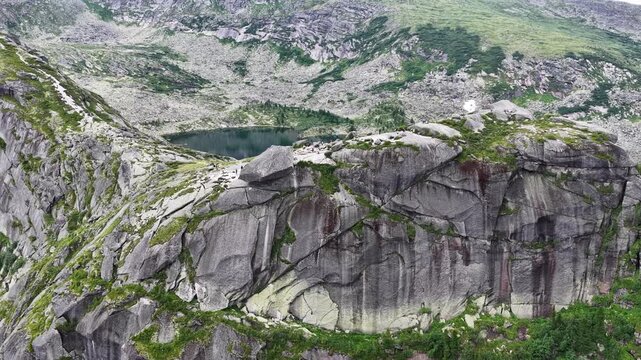 Breathtaking drone panoramic shot of remote mountain wilderness featuring sharp rocky ridges, dark green coniferous forest, and clear blue glacial lakes under shifting storm clouds.
