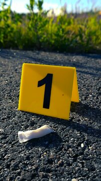 Yellow evidence marker with the number one next to a white nicotine pouch found on an asphalt road during a police investigation at a crime scene on a sunny day with a blurry background
