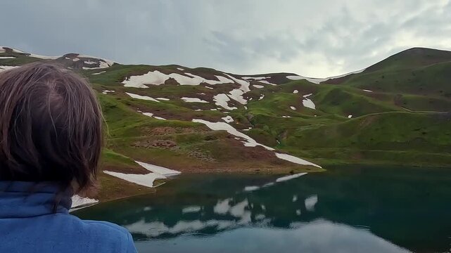 Tourist photographing Karagoli Lake over shoulder, capturing alpine water, snow patches, and green mountain slopes in Georgia for travel media, hiking routes, eco tourism, and nature destination