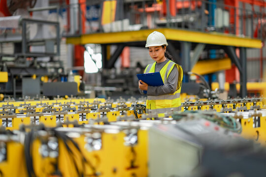 Female industrial engineer inspecting manufacturing equipment and recording production information on clipboard. Factory quality specialist performing routine inspection.