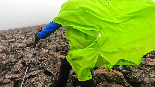 Hiker ascending rear view on Didi Abuli mountain in Georgia, pushing uphill through strong wind over loose scree with poles and green rain poncho for alpine hiking, weather risk, and outdoor travel