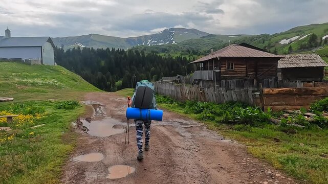 Backpacker walking in wide shot through mountain village in Georgia, carrying camping mat on muddy road with wooden houses and highland scenery for trekking route, rural tourism, and Caucasus travel