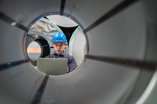 Industrial worker reviewing inventory information on laptop through steel coil. Digital warehouse management, manufacturing operations, material tracking, industrial technology, inventory control.