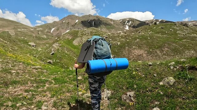 Hiker trekking rear view across Georgian alpine meadows, carrying large pack and camping mat toward mountain pass for hiking route media, wilderness travel, outdoor endurance, and Caucasus tourism