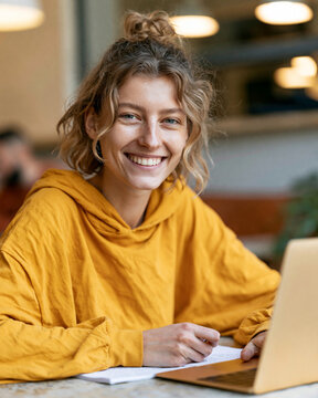 Warm smile lights up a cheerful young woman. She wears a soft yellow hoodie, sitting calmly. Her hair bounces gently, framing a joyful expression. Hands rest near a laptop, hinting at focused work