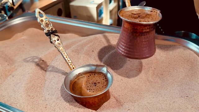 Close up of delicious oriental coffee boiling in copper pots placed in hot sand container. Professional barista hand holding ibrik handle making traditional turkish espresso beverage outdoors.