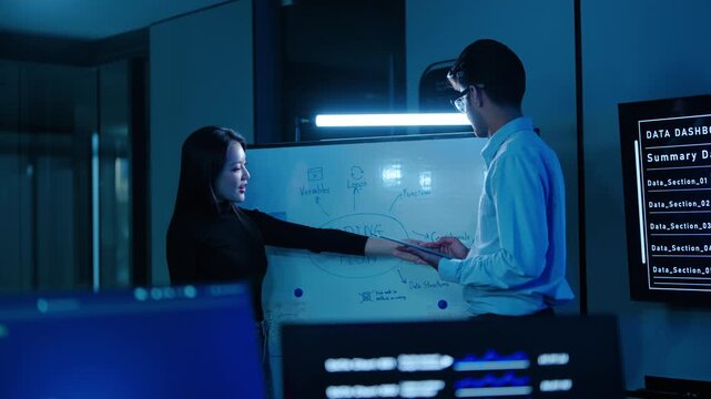Two Asian programmers brainstorming software architecture on a whiteboard in a dark high-tech office. Tech team discussing coding flow late at night. IT developers planning application logic.