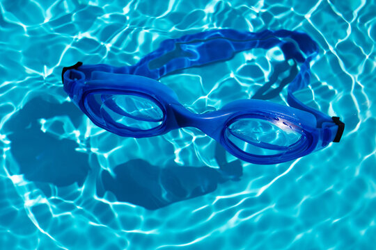 Swimming goggles on blue water sunlight surface in summer swimming pool. Underwater spectacles for diving, leisure and vacation.