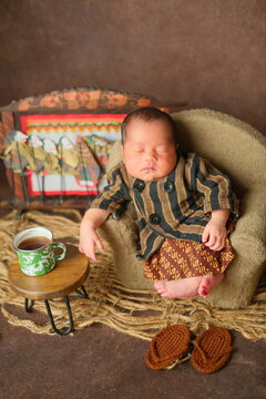 cute newborn baby boy sleeping in traditional javanese lurik clothes relaxing on mini sofa with tiny flip flops. from a different angle