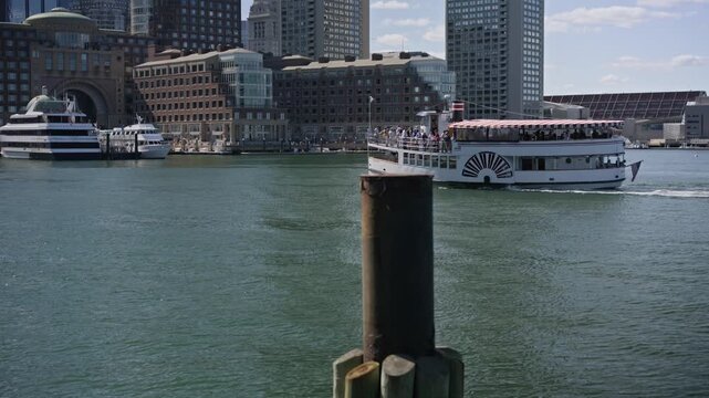 Cinematic view of a tourist boat cruising through Boston Harbor with the downtown Boston skyline in the background. Bright sunshine and calm waterfront waters showcase one of New Eng