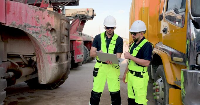 Two adult male logistics workers stand between industrial truck and container equipment, reviewing laptop schedule, cargo movement plan, safety briefing for freight yard task before dispatch