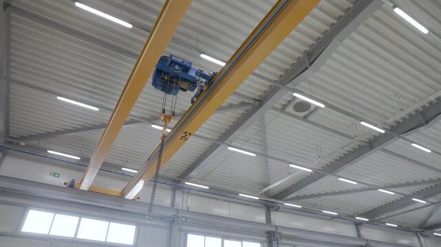 Yellow overhead crane with chain hoist mounted beneath a bright industrial ceiling in a modern factory