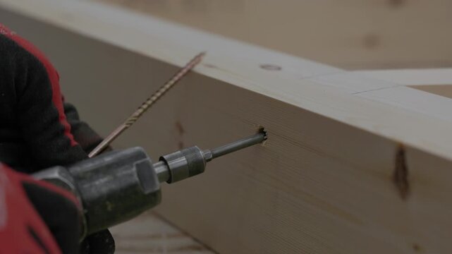 Close-up of a power drill driving a wood screw into a timber beam during frame assembly in a woodworking workshop. Shallow depth of field with copy space