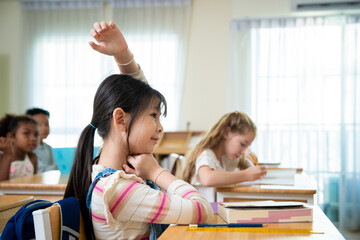 Obraz premium Cute Asian schoolgirl raising her hand to answer a question during a lesson in classroom. Smart elementary student raising hand for participation in class with classmates in background.