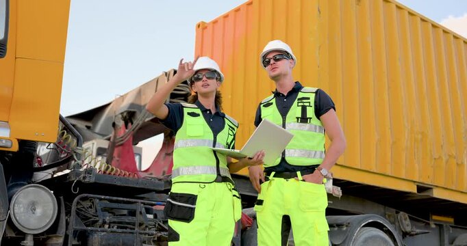 Adult Caucasian woman and man transport supervisor stand near yellow shipping container, signaling direction, checking laptop report, coordinating yard safety, cargo inspection, dispatch planning
