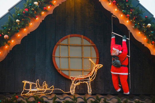 Jolly Santa climbing ladder on house decorated with festive garland, lights, glowing reindeer.