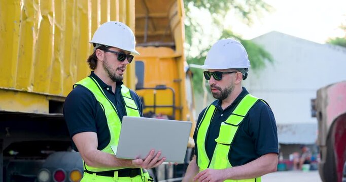 Adult male transport supervisor carries laptop near yellow container while colleague explains yard plan, coordinating cargo dispatch, freight schedule, safety compliance during site briefing