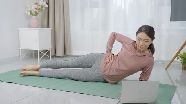 Young woman exercising at home on yoga mat following an online workout on a laptop in a bright living room, healthy lifestyle, stretching exercise and online digital fitness exercise concept