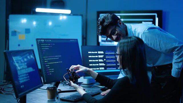 Young Asian IT engineers collaborate on a robotics project in a dark high tech lab. Man and woman program a small drone prototype, analyzing software code on multiple computer screens for innovation.