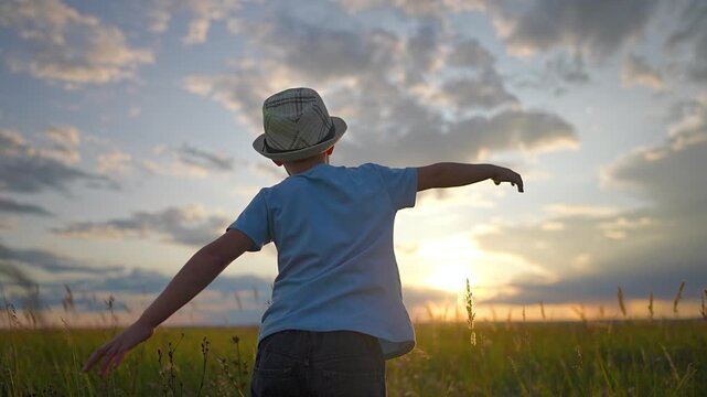 Happy boy stands in summer field with arms outstretched. Child dreams of flying like pilot at sunset. Kid plays airplane in green nature. Young boy wears hat and enjoys freedom. He looks at sky.
