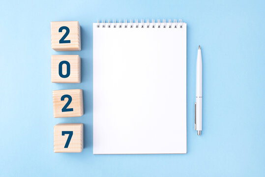 Wooden cubes with numbers for the New Year 2027 with a notebook and pen on a blue background