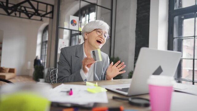 Portrait of successful mature businesswoman in modern office, smiling lady. Funny senior woman looking at laptop screen and rejoicing, investments and startup, dancing accountant or financial director