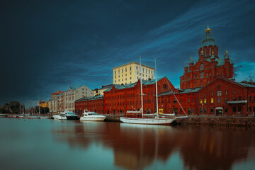 Obraz premium dramatic view of the historic red brick Uspenski Cathedral and surrounding buildings reflected in the calm waters of the harbor under a spectacular dark sunset sky.