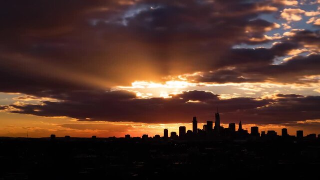 Sunset over city skyline with dramatic clouds and rays of sunlight illuminating buildings, creating a stunning visual transition from dusk to evening