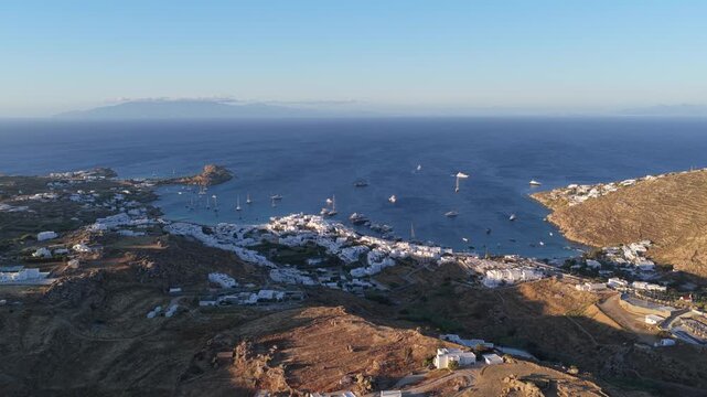 White washed architecture of Mykonos island in Greece overlooking Aegean Sea during sunset, showing beach, traditional houses, moored yachts, distant islands