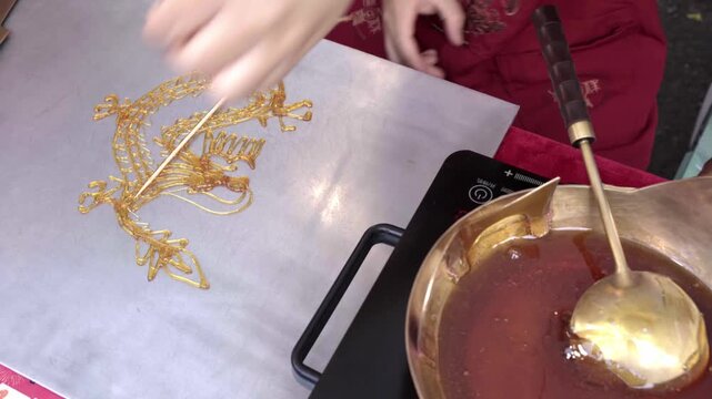 Close-up footage of a skilled artisan attaching a wooden skewer to a handcrafted Chinese sugar painting dragon (Tanghua) before carefully lifting it from the metal surface. Ttraditional confection