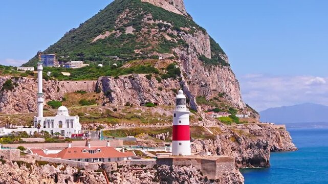 Drone footage panning sideways with telephoto compression past Gibraltar's red and white lighthouse and white mosque, stacking sheer limestone cliffs, blue strait, and distant African coastline.