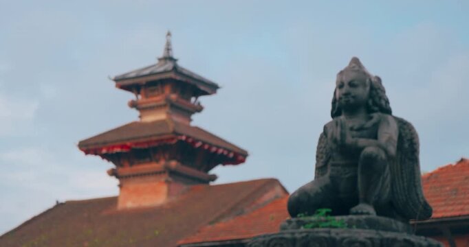 Bhaktapur Durbar Square, Nepal: A stone-carved Garuda sculpture kneeling with a traditional tiered pagoda-style temple blurred in the background at UNESCO heritage site.