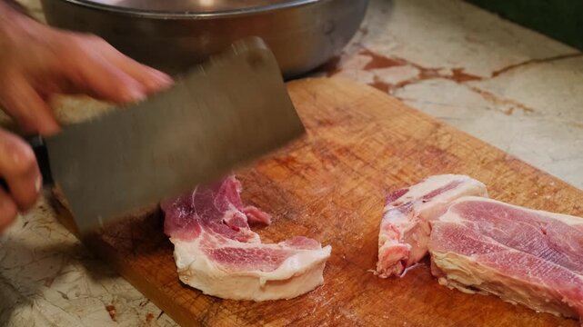 Chef chopping raw pork into thick slices with a traditional meat cleaver on a wooden cutting board. Authentic barbecue preparation highlighting fresh ingredients and culinary craftsmanship.