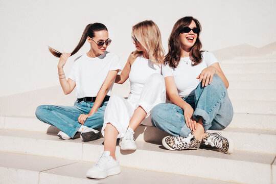 Three young beautiful smiling hipster female in trendy summer t-shirt  and jeans clothes. Sexy carefree women posing in street. Positive models having fun, sit at stairs. In sunny day, sunglasses