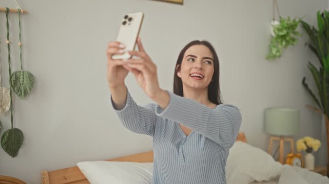 Woman holding smartphone and extending arms for a selfie, puckered lips and smiling expression on bed in a bedroom; playful confidence.