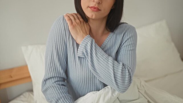 Woman with hand on shoulder sitting on bed, touching collarbone and shoulder near pendant necklace, wearing ribbed sweater with blanket in building; gentle discomfort.